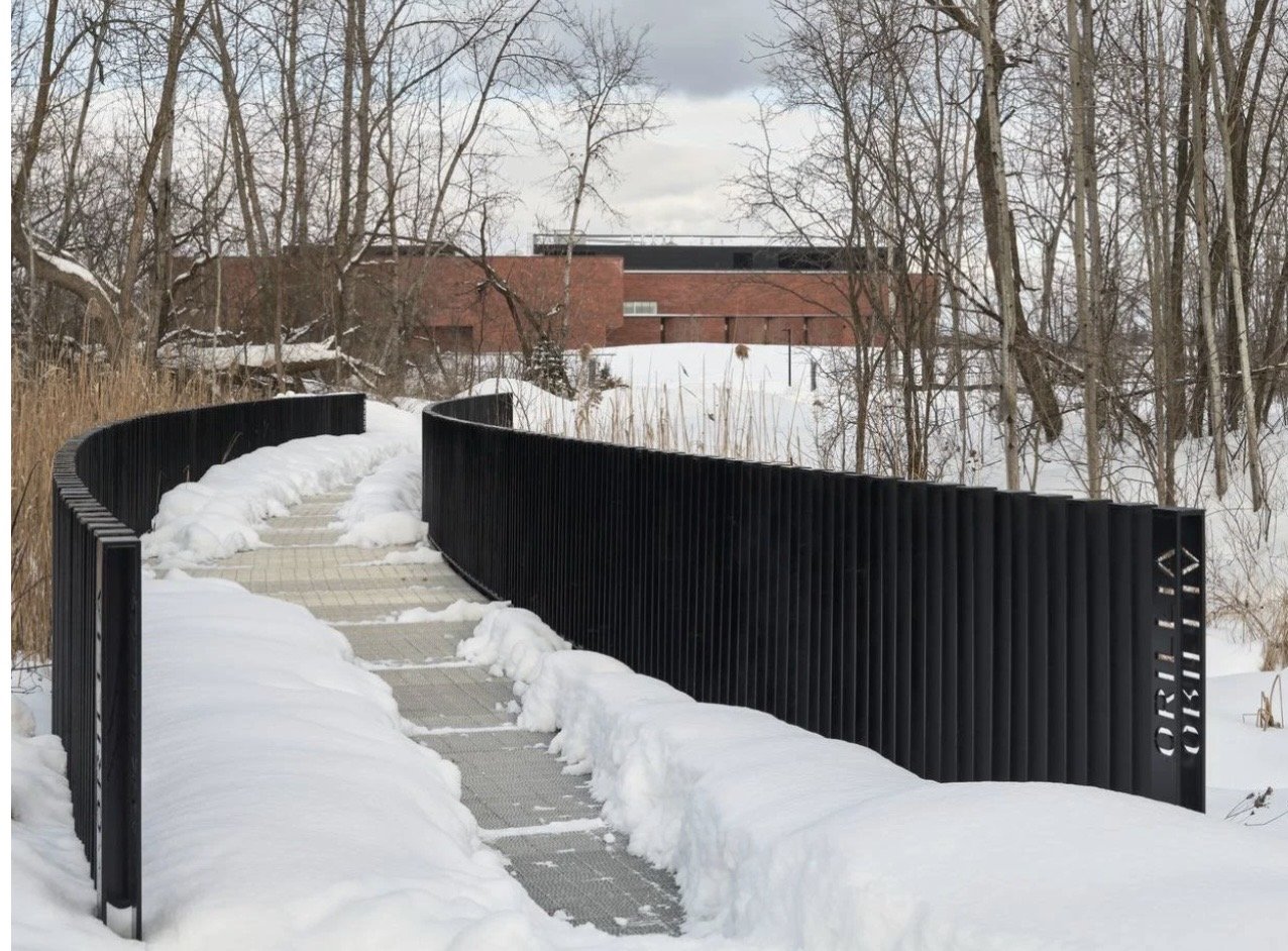 Orillia Recreation and Aquatic Centre – Curved pedestrian bridge with black vertical-slat railings in snow, the brick recreation centre in the background